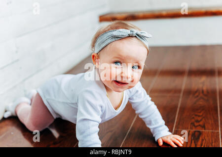 Little crawling baby girl one year old siting on floor in bright light living room near window smiling and laughing. Happy toddler kid playing at home Stock Photo