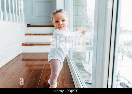 Little crawling baby girl one year old siting on floor in bright light living room near window smiling and laughing. Happy toddler kid playing at home Stock Photo