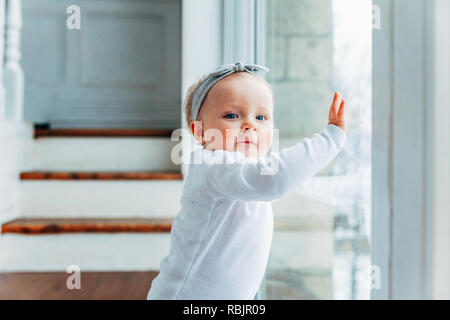 Little crawling baby girl one year old siting on floor in bright light living room near window smiling and laughing. Happy toddler kid playing at home Stock Photo