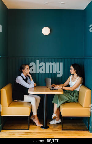 Two young women from a startup have a working meeting in a trendy booth in their office during the day. One is Caucasian and the other Asian Chinese. Stock Photo