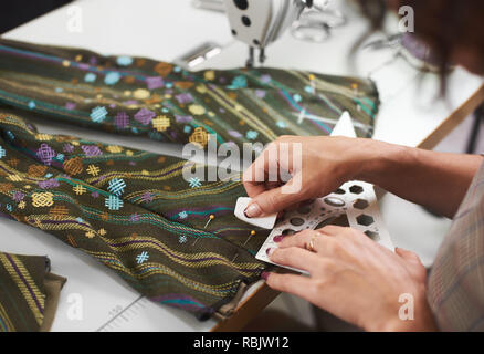 Tailor workplace: close-up of woman hands making marking on garment details using chalk and ruler before sewing on electrical sewing machine. Handmade Stock Photo