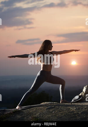 Slim attractive long-haired girl with stretched arms in standing yoga pose outdoors on big boulder on background of cloudy beautiful foggy pink sky an Stock Photo