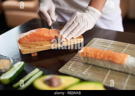 Close-up view of process of preparing delicious rolling sushi in restaurant. Female hands in disposable gloves slicing salmon. Stock Photo
