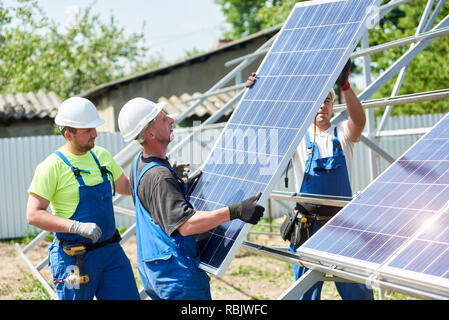 Team of three young technicians in protective helmets mounting heavy solar photo voltaic panel on metal platform in back yard of village house. Cheap  Stock Photo
