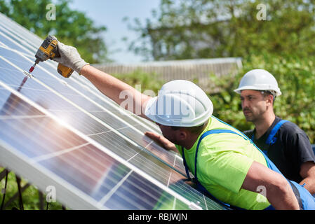 Professional two technicians working with screwdriver connecting solar photo voltaic panel to metal platform on summer rural landscape background. Sta Stock Photo
