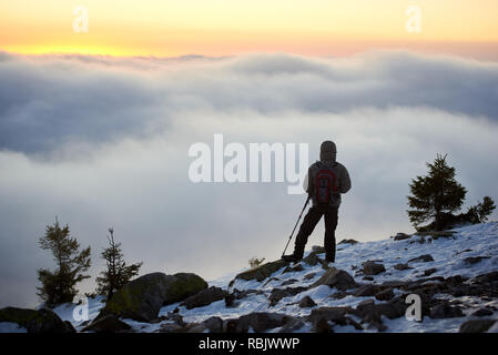 Back view of tourist hiker with backpack and hiking poles on the top of rocky mountain, enjoying view of valley covered with white clouds stretching t Stock Photo
