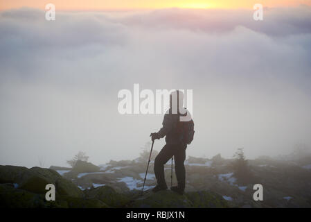 Silhouette of tourist hiker with backpack and trekking poles on mountain big rocks enjoying beautiful view of clouds down below in valley and bright y Stock Photo