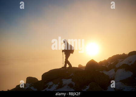 Silhouette of tourist hiker with backpack and hiking sticks on rocky snowy mountain on copy space background of bright white raising sun and misty ora Stock Photo