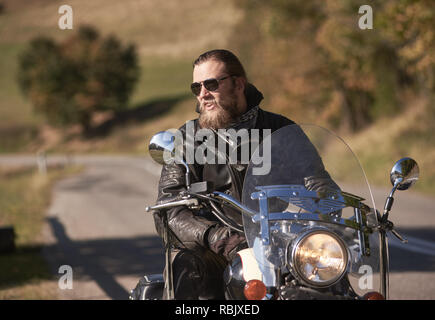 Portrait of handsome happy smiling bearded biker in black leather jacket and sunglasses on modern motorcycle on country roadside on blurred background Stock Photo