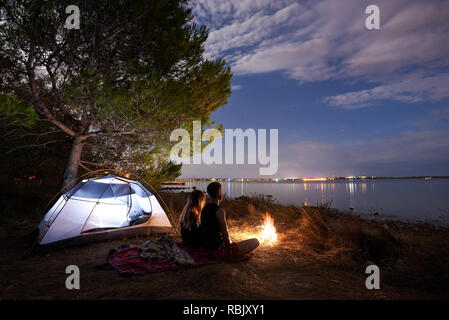 Camping night on sea shore. Young couple, bearded man and pretty woman backpackers having a rest near tourist tent by campfire under blue cloudy eveni Stock Photo