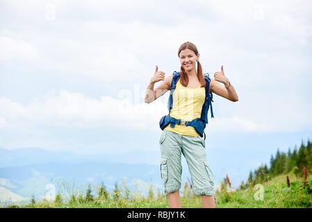 Portrait of attractive smiling woman tourist hiking mountain trail, walking on grassy hill, wearing backpack, using smiling to the camera and showing  Stock Photo