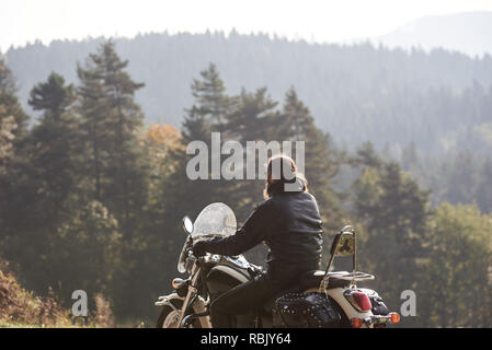 Back view of bearded biker with long hair in black leather jacket sitting on cruiser motorcycle on blurred background of spruce tall trees and woody hills on horizon. Active lifestyle and traveling. Stock Photo