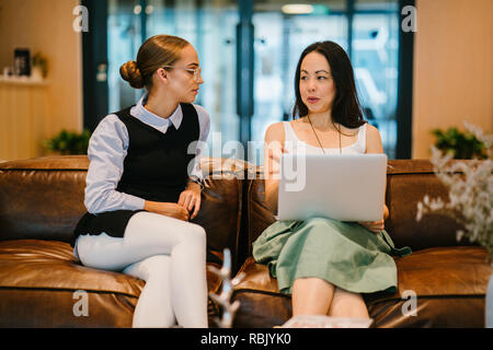 Two colleagues in a diverse workplace have a meeting together on a couch in a trendy office. One woman is Caucasian and the other Asian Stock Photo