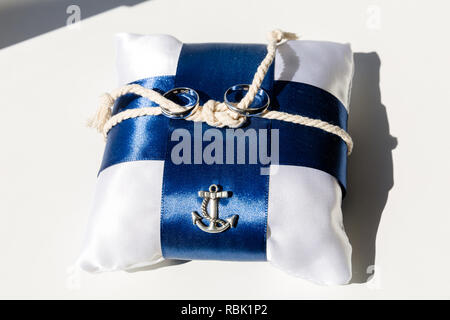 A pair of wedding rings on a white pillow with blue ribbons. In the middle of the pillow is a rope tied up with a sea knot. On the side of the pillow is a metal decoration in the shape of a sea anchor Stock Photo