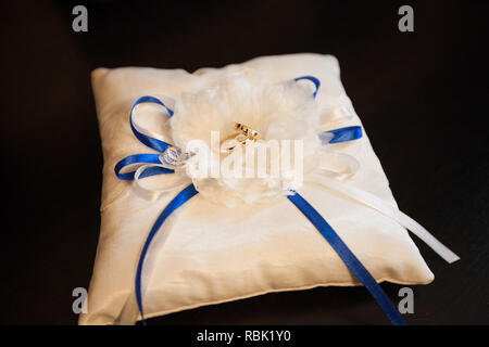 A pair of wedding rings on a white pillow. In the center of the pillow is a bow made of white and blue ribbons. Pillow on a black background. Stock Photo