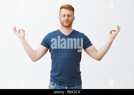 Calm down, peace inside. Relaxed good-looking european guy with red hair and bristle, closing eyes, standing with hands spread aside with zen gesture, meditation, relieving from stress with yoga Stock Photo