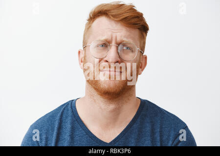 Handsome gloomy redhead male with beard frowning and pursing lips looking right with regret and disappointment, standing over gray background in casual clothes and glasses Stock Photo