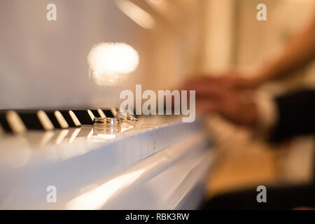 A pair of wedding rings are on the keys of the white piano. In the background in the blur distinguishable hands of the bride and groom Stock Photo