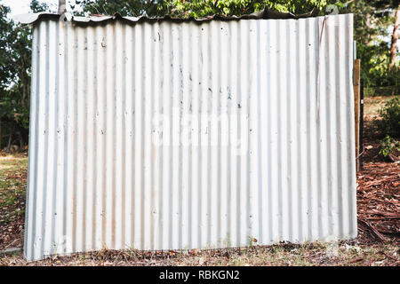 Side of a small rustic, bright looking corrugated iron shed with a shaded back yard scene behind. Stock Photo