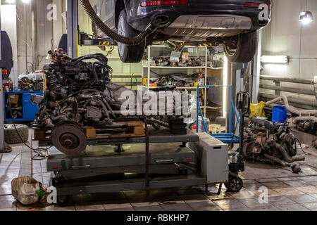 An SUV car raised on a lift for repair and under it a detached engine suspended on a gray lifting table in a vehicle repair workshop with a lot of lig Stock Photo