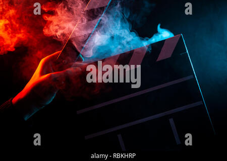 Close-up on an open clapper in hand before starting shooting a film with multi-colored smoke around with red and blue backlighting on a black isolated Stock Photo