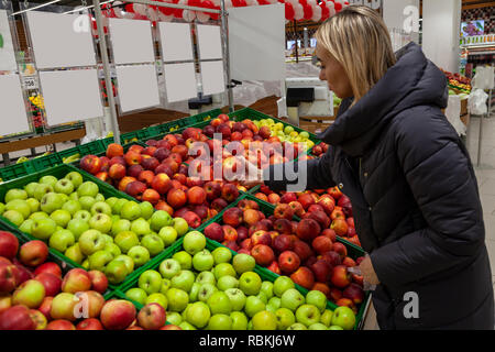 A blond woman in a supermarket buys apples from the beautifully laid out rows on the shelves, stopping her choice on red, holds it in her hand. Stock Photo