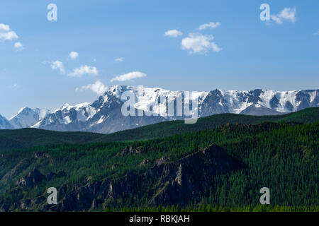 The picturesque landscape of the valley of hills covered with green grass and trees in front of the stone ridge of snow-capped peaks on which lies whi Stock Photo