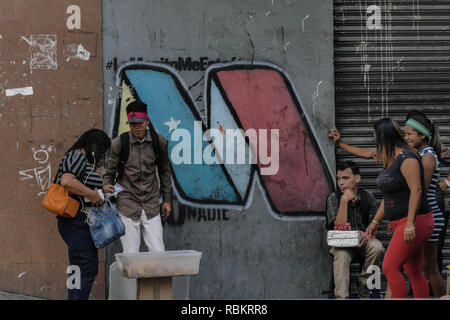 Caracas, Miranda, Venezuela. 10th Jan, 2019. A graffitti with the M of Maduro is seen on a wall, in front of the graffitti illegal vendors sells snacks and cigara in the street.Venezuelan President Nicolas Maduro started a second term, defying critics in the United States and Latin America who called him an illegitimate usurper of a nation where economic chaos has wrought a humanitarian crisis. The National Assembly calls the Maduro government a ''dictatorship and usurpation of power.'' Currently Venezuela is experiencing its worst economic crisis with an inflation rate of over 1,000,000% Stock Photo