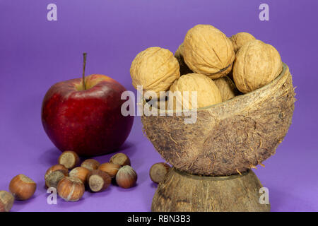 Whole common walnuts with shells in natural coconut shell cup, pile of whole hazelnuts and red apple on purple background Stock Photo