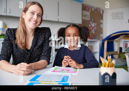 Young female primary school teacher and schoolgirl sitting at a table, working one on one in a classroom, smiling to camera, front view, close up Stock Photo
