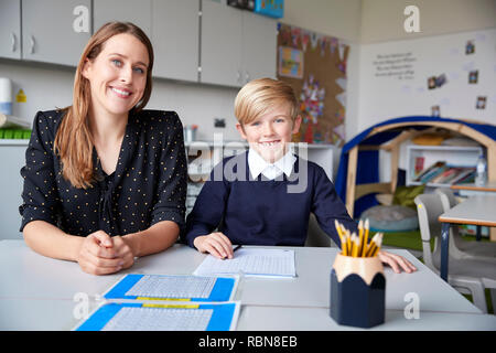 Young female primary school teacher and schoolboy sitting at a table, working one on one in a classroom, smiling to camera, front view, close up Stock Photo