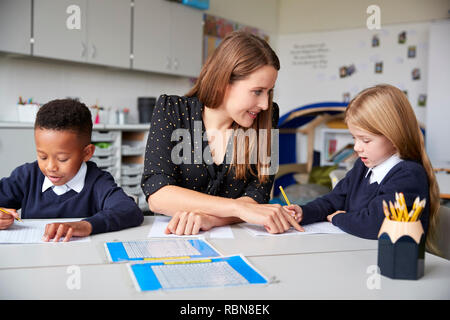 Female school teacher sitting between two primary school kids at a table in a classroom, helping a girl with her work, close up Stock Photo