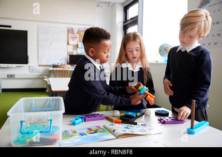 A girl and two boys standing at a table in a primary school classroom working together with toy construction blocks, close up Stock Photo