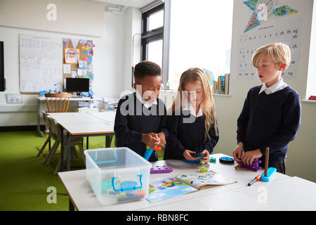 A girl and two boys standing at a table in a primary school classroom working together with toy construction blocks Stock Photo