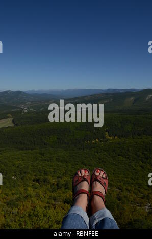 Female legs in trekking shoes over a huge precipice against a background of green forest and blue sky. Crimea. Ukraine Stock Photo