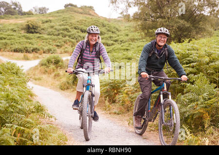 Senior couple riding mountain bikes in a country lane during a camping holiday smiling, front view Stock Photo