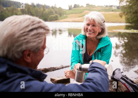 Senior couple by a lake, man pouring coffee to his wife’s cup, over shoulder view, Lake District, UK Stock Photo