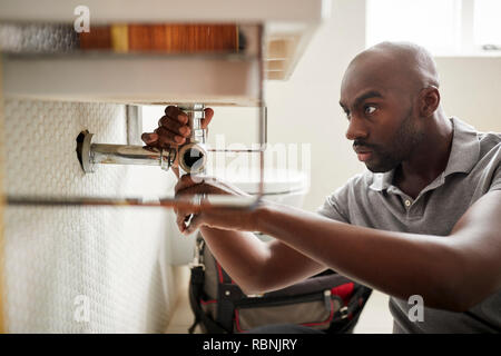 Young black male plumber sitting on the floor fixing a bathroom sink, close up Stock Photo
