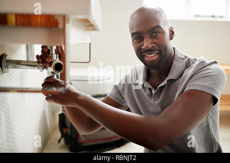 Young black male plumber sitting on the floor fixing a bathroom sink, looking to camera, close up Stock Photo