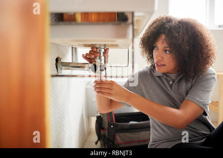 Young black female plumber sitting on the floor fixing a bathroom sink, seen from doorway Stock Photo
