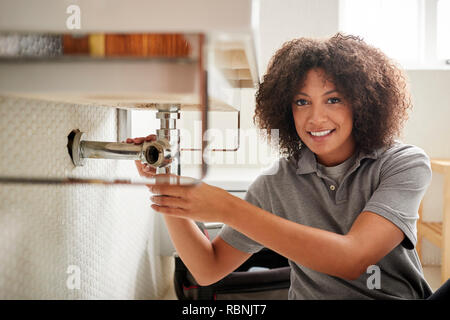 Young black female plumber sitting on the floor fixing a bathroom sink, looking to camera Stock Photo