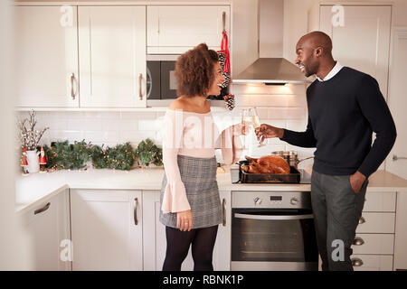 Young adult mixed race couple make a toast with champagne in their kitchen while preparing Christmas dinner, side view Stock Photo