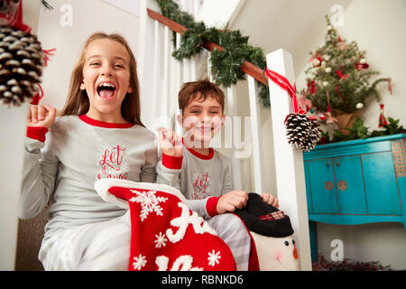 Portrait Of Two Excited Children Wearing Pajamas Sitting On Stairs Holding Stockings On Christmas Morning Stock Photo