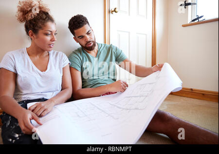 Couple Sitting On Floor Looking At Plans In Empty Room Of New Home Stock Photo