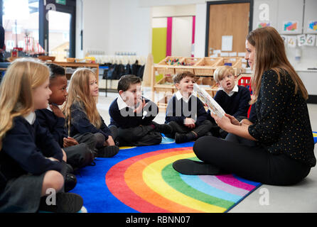 Primary school kids sitting on the floor in class listening to their female teacher reading a book to them, side view Stock Photo