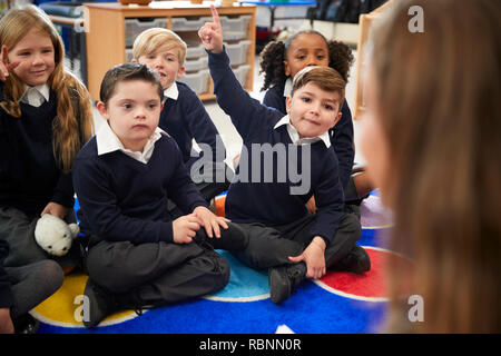 View over female teacher's shoulder of primary school kids sitting on the floor in the classroom raising hands to answer a question Stock Photo