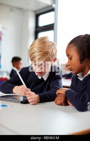 Two primary school kids using a tablet computer and stylus sitting together at desk in a classroom, vertical Stock Photo