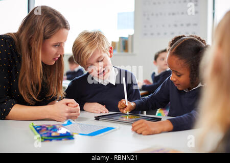Female school teacher helping two kids using a tablet computer at desk in a primary school classroom, close up Stock Photo