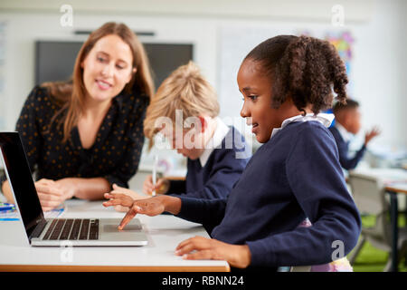 A female school teacher sitting at a desk helping a boy and girl using a laptop computer in primary school class, side view