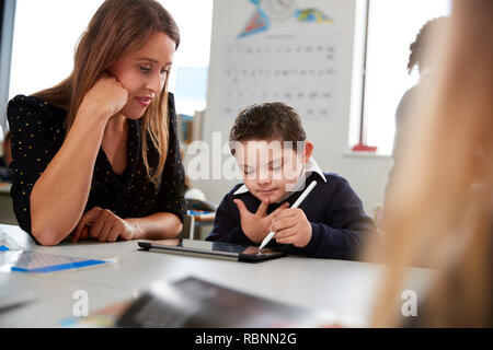 Young female teacher working with a Down syndrome schoolboy sitting at desk in a primary school classroom, selective focus Stock Photo